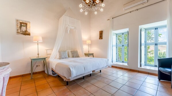 Une chambre à coucher avec un lit à baldaquin, deux tables de chevet avec des lampes, une grande fenêtre donnant sur un paysage verdoyant et un lustre.