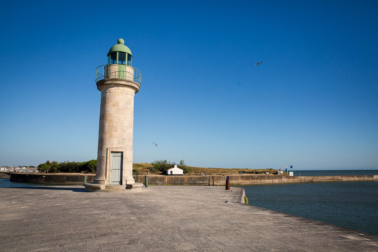 A lighthouse with a green dome, situated on a paved area near a body of water.