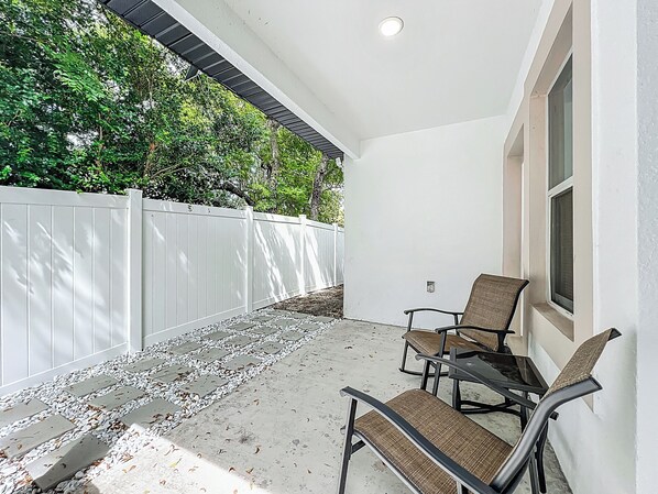 A covered patio with a white fence, a concrete pathway, and two chairs.