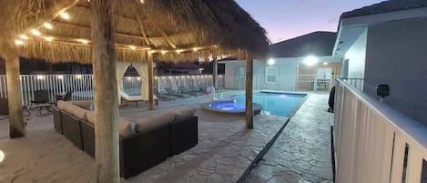 An outdoor pool area with a thatched roof, lounge chairs, and a hot tub.