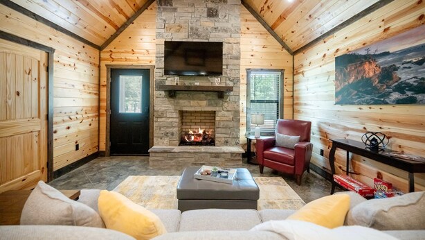 A cozy log cabin interior with a stone fireplace, a comfortable seating area, and a mounted TV. - Couples Escape w/ Hot Tub•Firepit •Trail Access (Broken Bow)