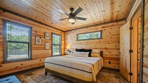 A wooden cabin bedroom with a bed, a ceiling fan, and large windows. - Couples Escape w/ Hot Tub•Firepit •Trail Access (Broken Bow)