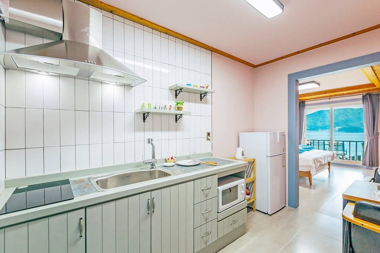 A modern kitchen with a built-in stove, microwave, and sink. There are shelves above the counter and a bed in the adjacent room.