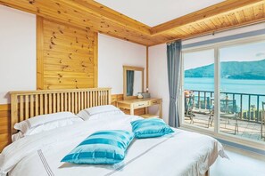 A bedroom with a wooden ceiling, a large bed with white and blue bedding, a wooden nightstand, a mirror, and a balcony with a view of a lake and mountains. - SINBIBADA PENSION (Geoje)