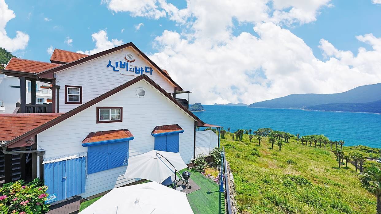 A coastal building with a blue and white exterior, a sign in a foreign language, and a view of the sea and distant mountains.
