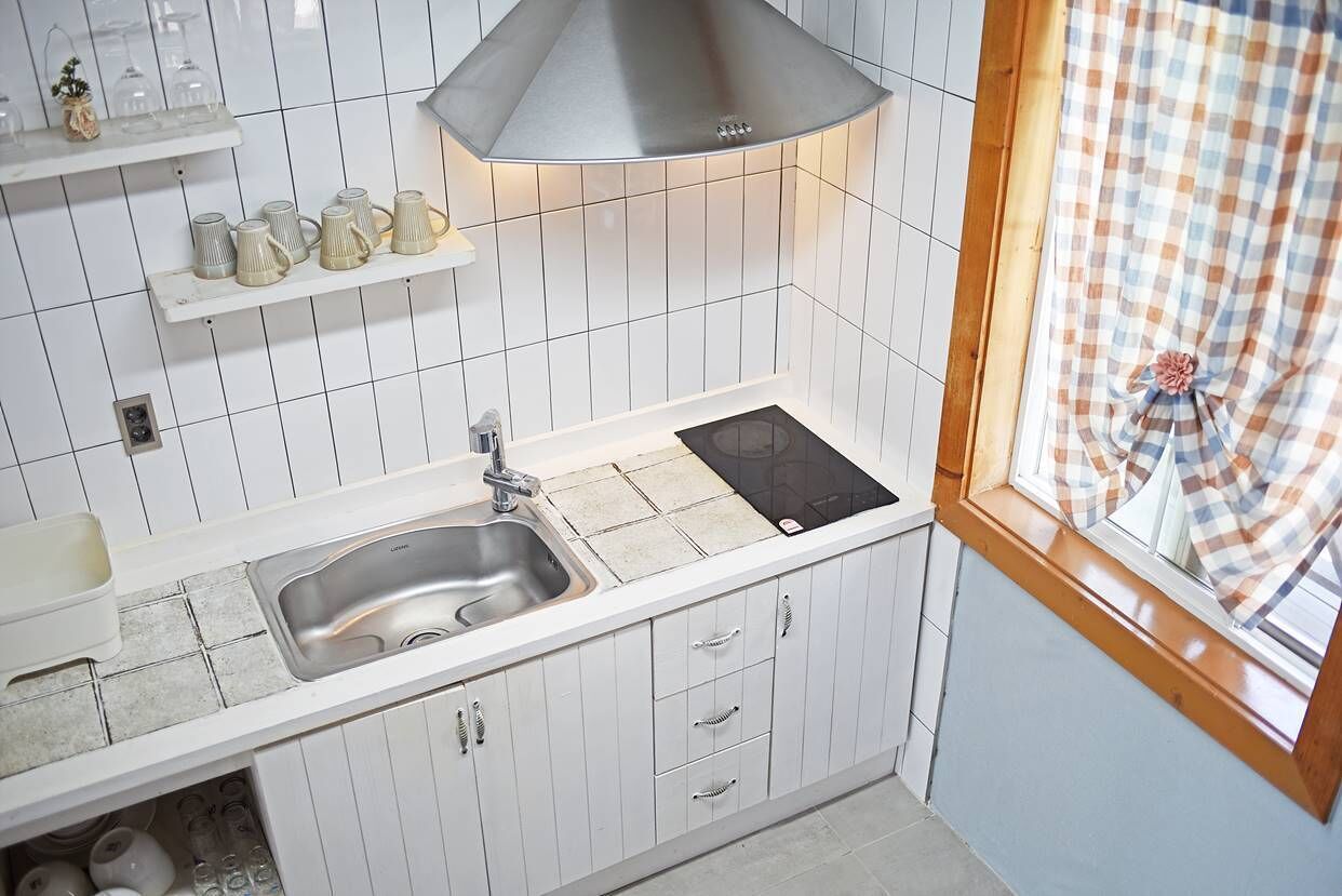 Una cocina con armarios blancos, un lavamanos de acero inoxidable, una placa de inducción negra y una ventana con cortina a cuadros.