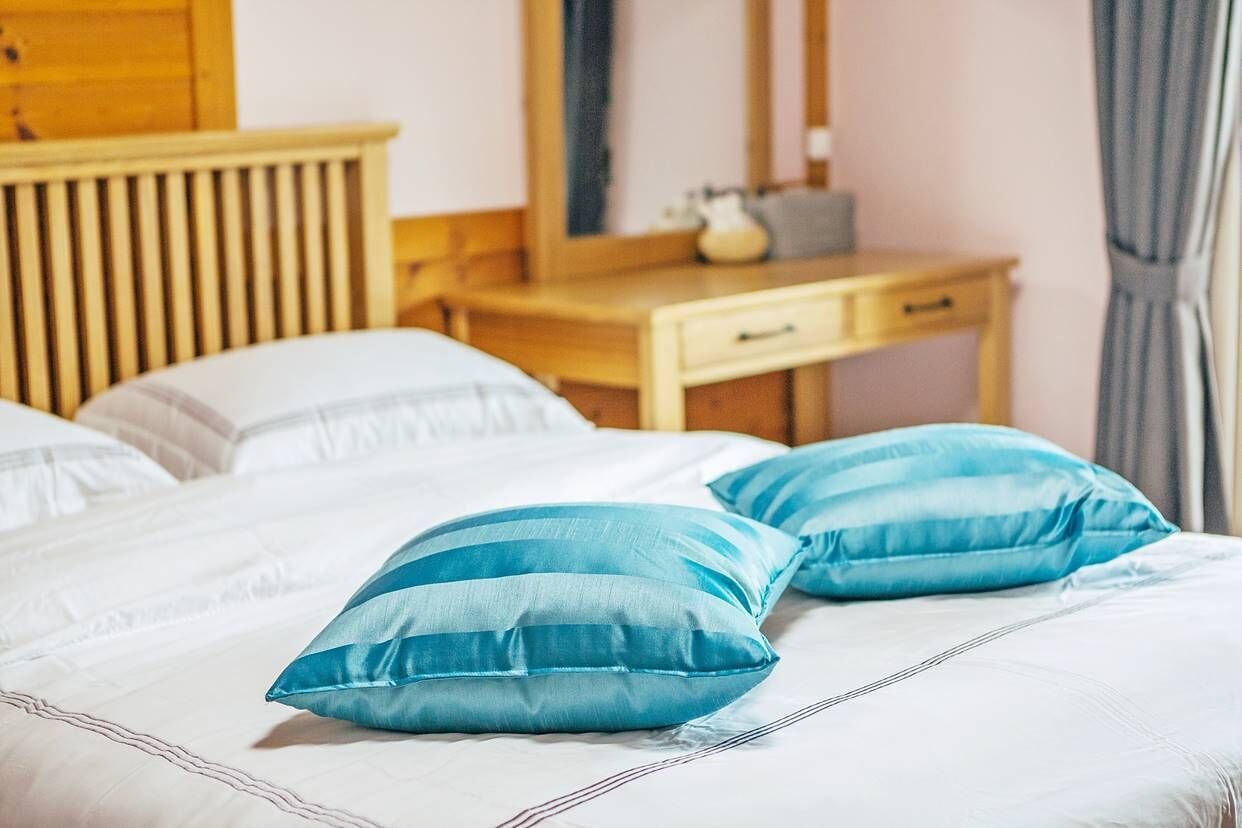 A neatly made bed with white linens and two blue pillows, a wooden nightstand with a lamp, and a wooden dresser with a mirror.