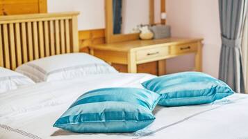 Una cama bien tendida con sábanas blancas y dos almohadas azules, una mesita de noche de madera con una lámpara, y un aparador de madera con un espejo.