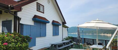 Un edificio blanco con contraventanas azules y un letrero en idioma extranjero. Hay mesas y sillas de picnic afuera, y un paraguas grande que proporciona sombra.