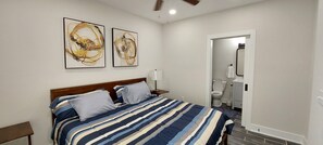 A bedroom with a bed, two paintings, a nightstand, and a bathroom visible through an open door.