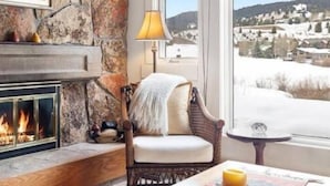 A cozy room with a stone fireplace, a comfortable chair, a table with a candle, and a view of a snowy landscape. - Silver Moose Condo (Gallatin Gateway)