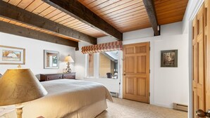 A bedroom with a large bed, wooden ceiling beams, a window with a view, and a door. - Silver Moose Condo (Gallatin Gateway)