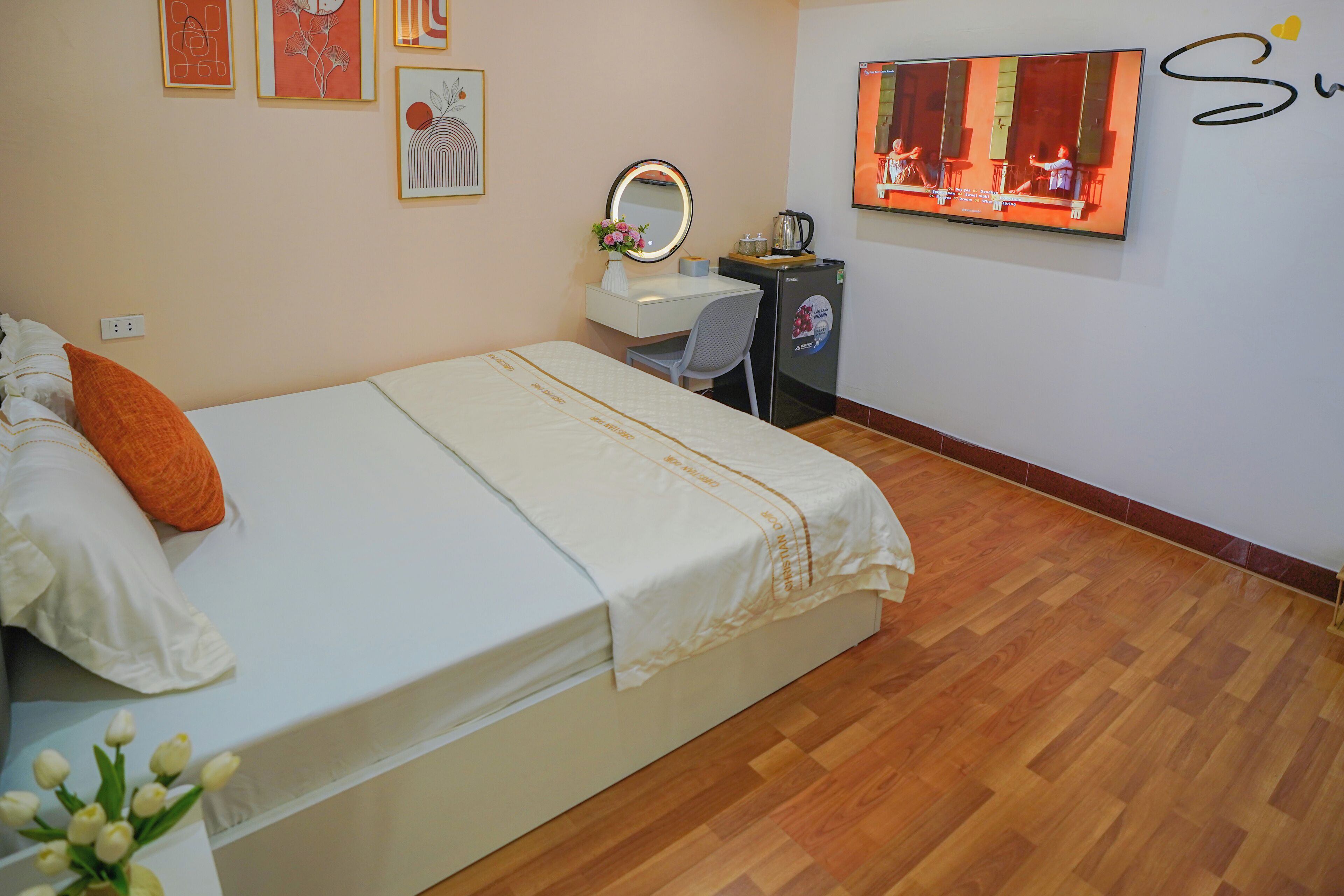 Habitación de hotel con cama, escritorio con silla, espejo, televisor y refrigerador pequeño.