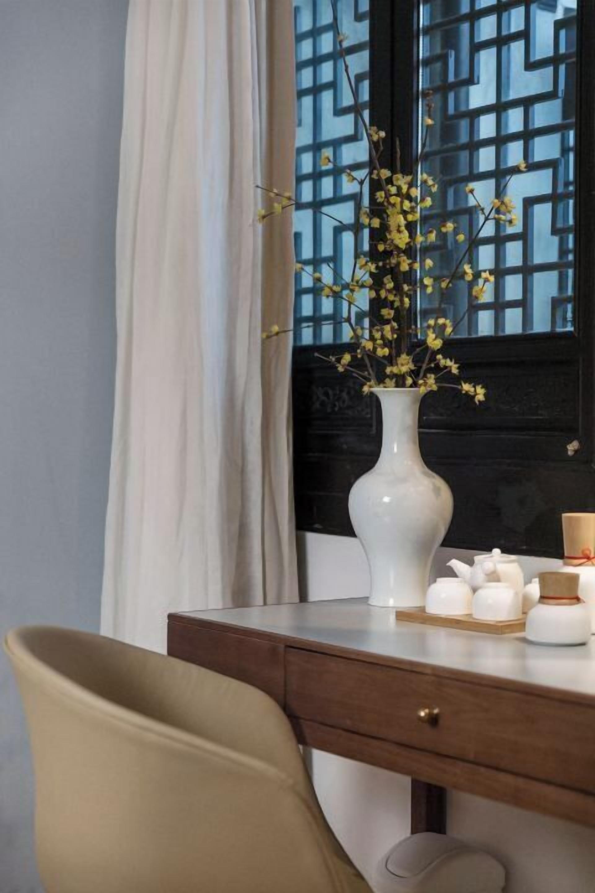 A room with a desk, a vase with yellow flowers, and a window with a geometric pattern.