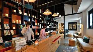 A modern reception area with a wooden counter, bookshelves, and a seating area with a sofa and round tables. - Blssom House Nanxun Huzhou  (Huzhou)
