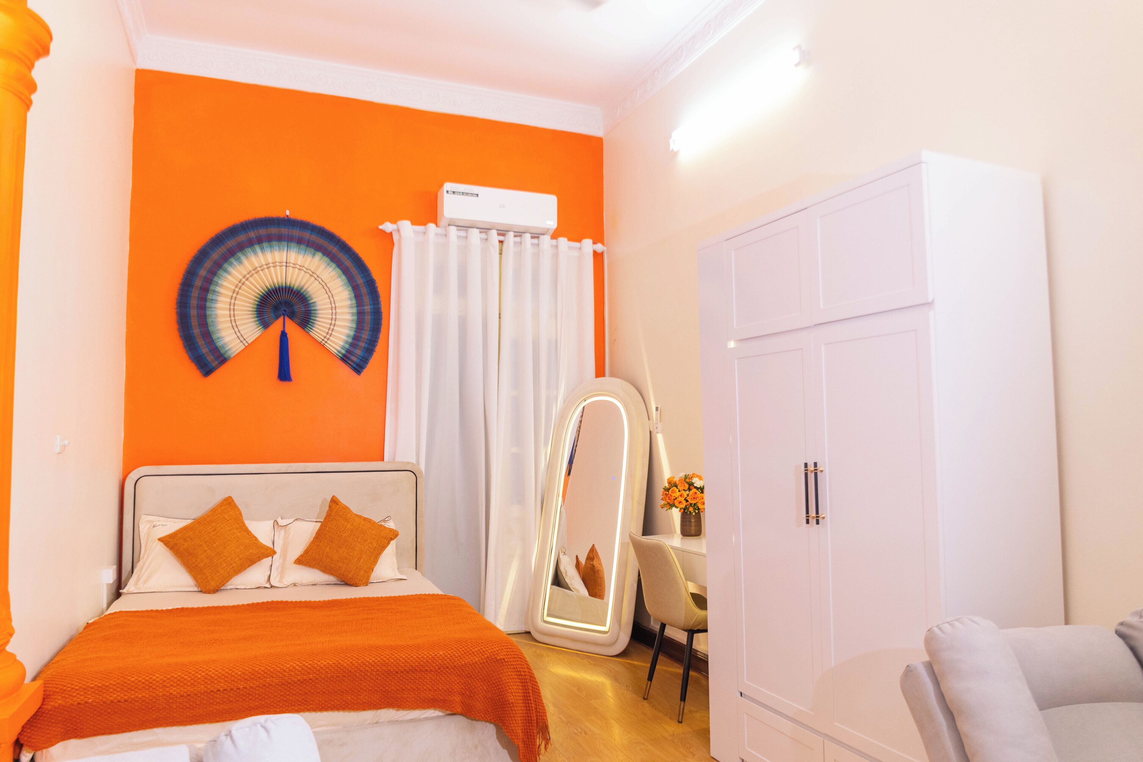 A bedroom with a bed, a white wardrobe, a desk with a chair, and an air conditioning unit on the wall.