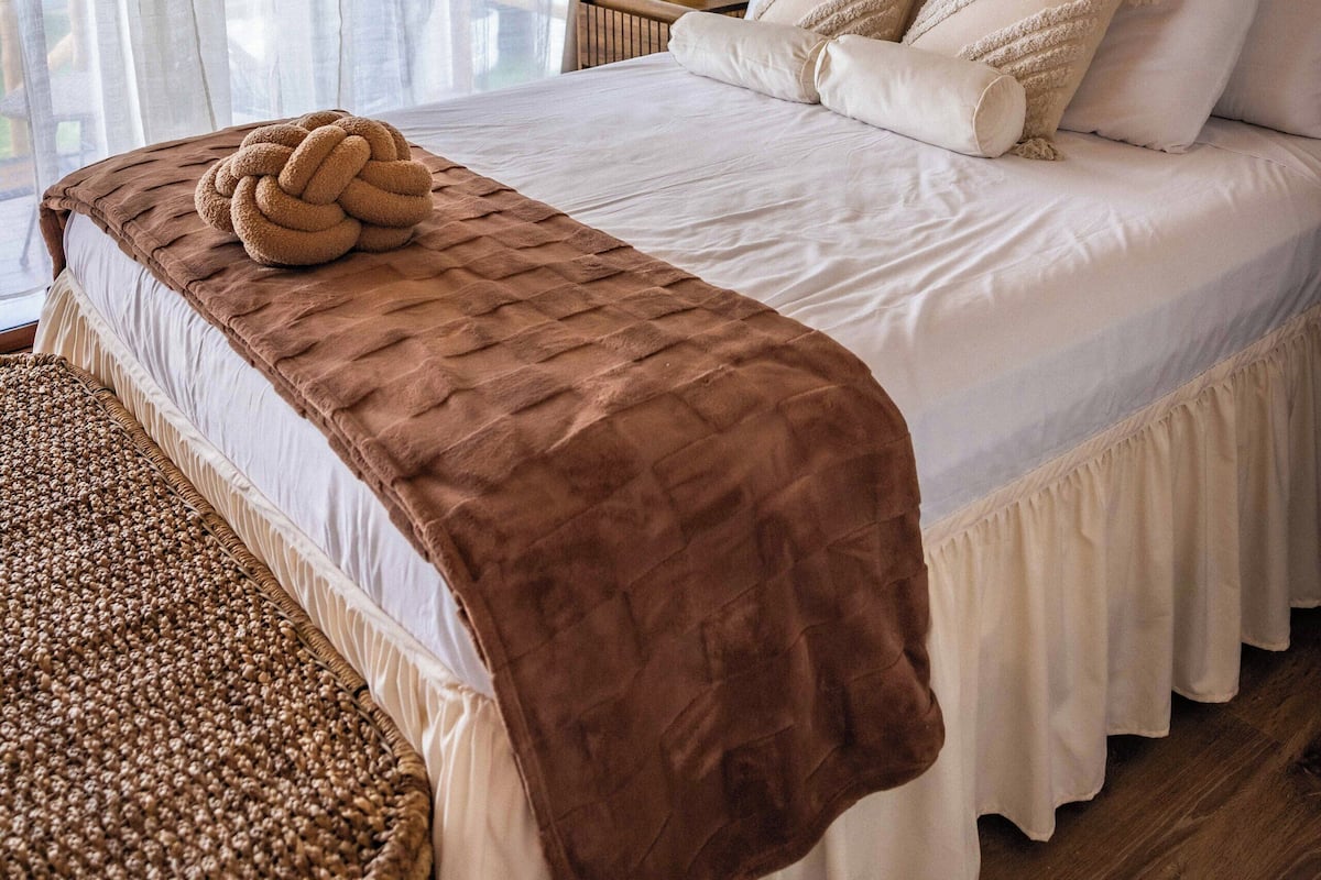 Een netjes opgemaakt bed met een bruine plaid, een geweven bankje en een hoofdbord met houten afwerking.