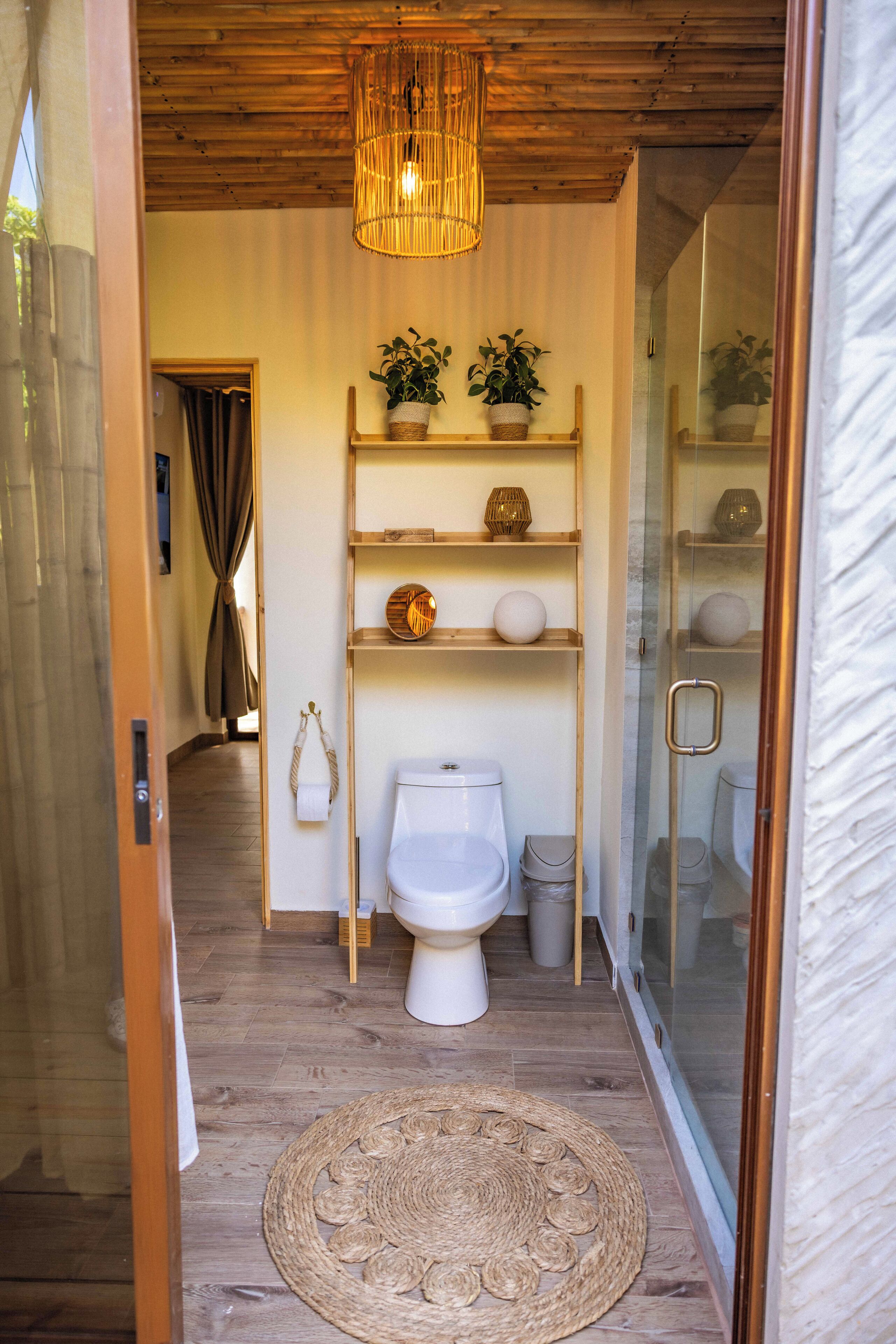 A bathroom with a wooden ceiling, a woven rug, a toilet, a shelf with decorative items, and a glass shower enclosure.