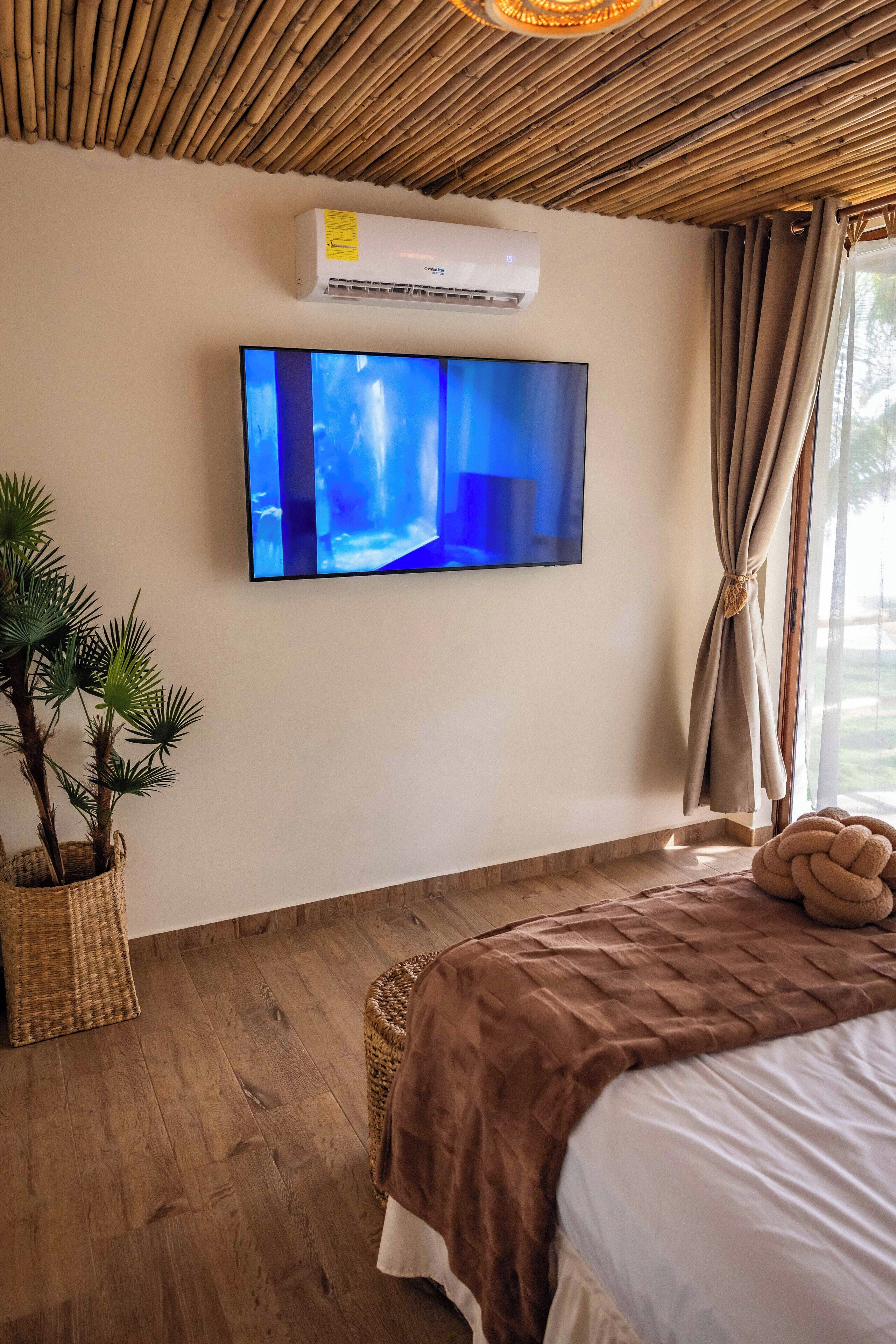 A bedroom with a large bed, a flat-screen TV mounted on the wall, a woven basket with plants, and a ceiling fan.