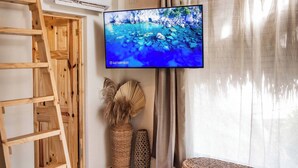 Huoneessa on puinen katto, seinälle asennettu televisio, punottu penkki, tikapuut ja punottu jalkarahi.