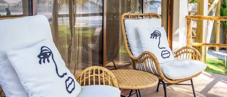Un balcón con sillas de mimbre, una mesa de madera y vistas a palmeras y una playa.