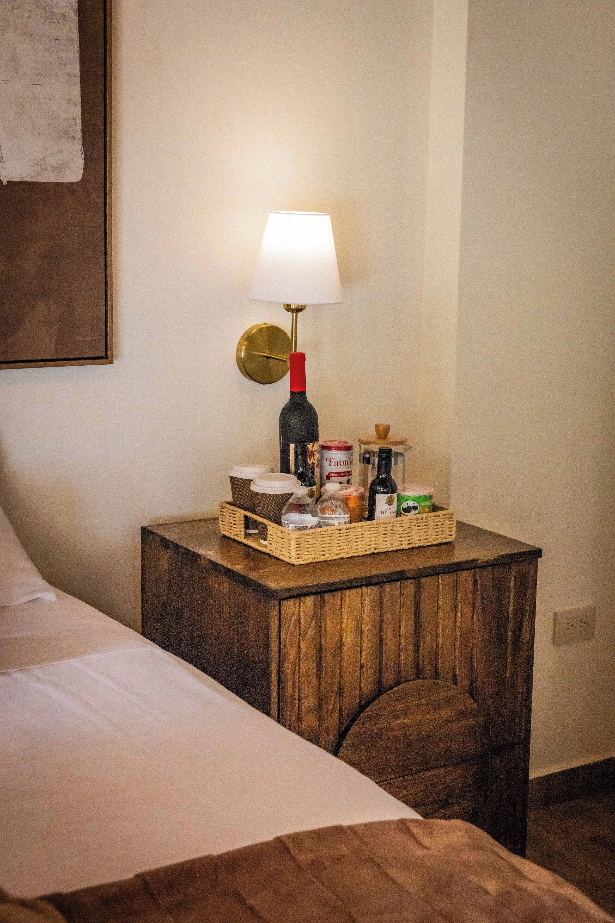 A neatly made bed with a white sheet, a wooden nightstand with a lamp, and a basket containing various items like a bottle, jars, and a candle.