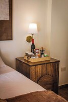 Una cama bien hecha con sábana blanca, una mesita de noche de madera con una lámpara, y una cesta con varios objetos como una botella, tarros y una vela.