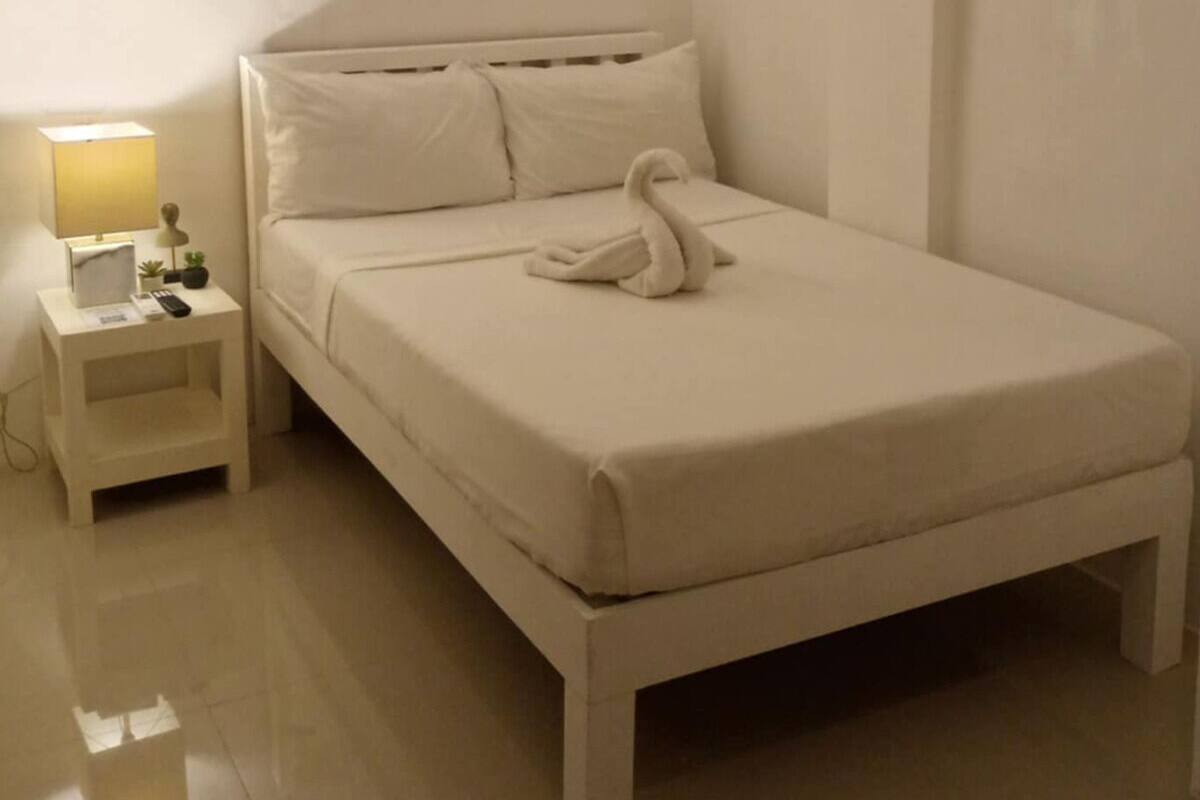 A single-bed room with a nightstand, a lamp, a remote control, and a swan-shaped towel.