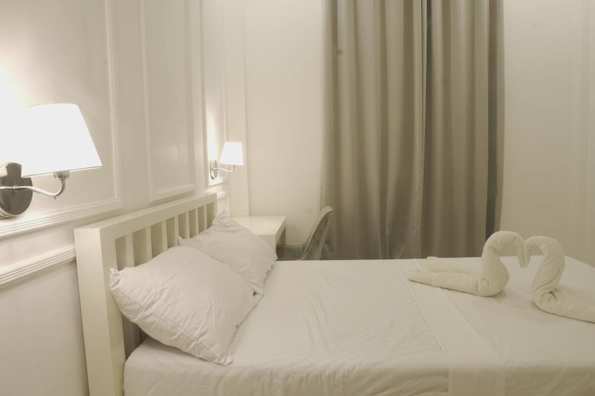 A hotel room with a white bed, two pillows, a bedside lamp, a chair, and two swan-shaped towels.