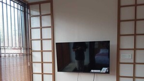 Ein Zimmer mit an der Wand befestigtem Fernseher, einem Holzschrank, einem Fenster mit Jalousien und einem kleinen Holztisch.
