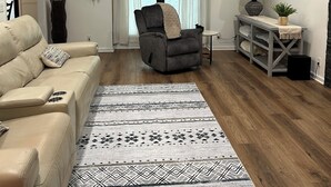 Un salón con un sofá color beige, un sillón gris, un tapete estampado, un ventilador de techo y una ventana con cortinas.
