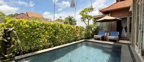A swimming pool with a stone statue, a hedge, and two lounge chairs with an umbrella.