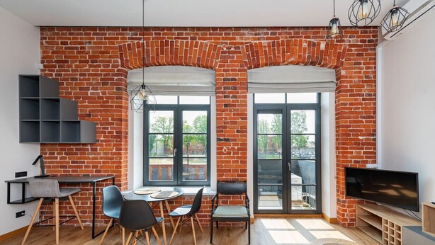 Una sala de estar moderna con paredes de ladrillo a la vista, un comedor con una mesa redonda y sillas, un televisor sobre un mueble de madera, y un balcón con vistas.