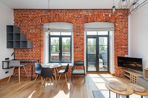 A modern living room with exposed brick walls, a dining area with a round table and chairs, a TV on a wooden stand, and a balcony with a view.