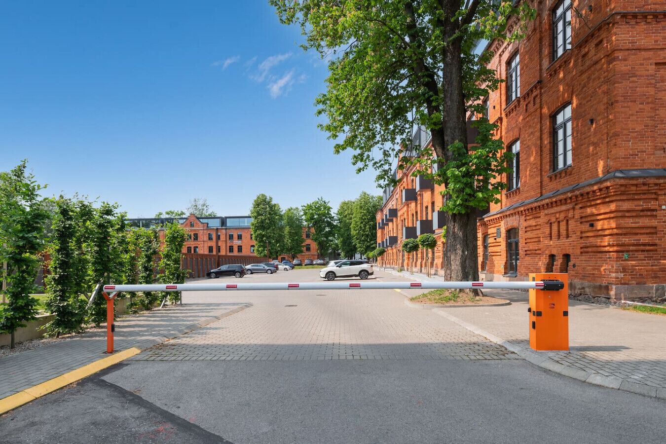 Une entrée fermée par un portail donnant sur un ensemble d’immeubles à l’architecture en briques, avec un ciel dégagé et des voitures garées.