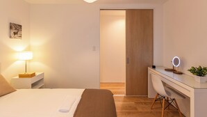 A bedroom with a bed, bedside table, lamp, desk, chair, and a potted plant.