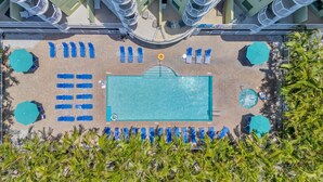 Vue aérienne d’un espace piscine doté de nombreuses chaises longues et de parasols, entouré de palmiers et d’un bâtiment avec des balcons.