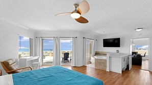 Une chambre spacieuse avec un grand lit, un ventilateur de plafond, une vue sur l’océan, un balcon avec une chaise et une petite kitchenette.