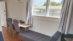 Habitación pequeña y funcional con sofá, una mesita y dos sillas. Se ve una ventana con cortinas y una puerta a la izquierda.