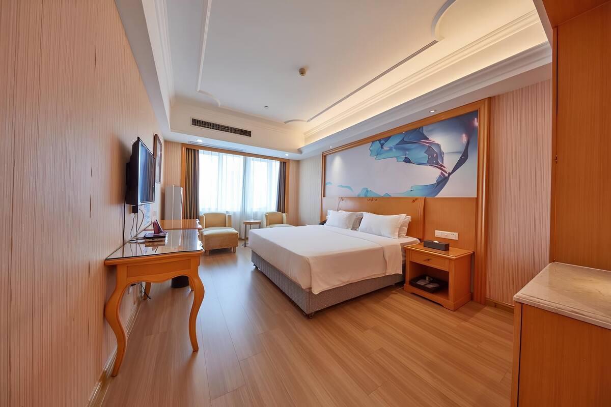 Ein Hotelzimmer mit einem großen Bett, einem Schreibtisch, einem Flachbildfernseher, einem Sessel, einem kleinen Tisch und einem Nachttisch.