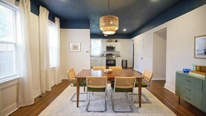 Een eetkamer met een houten tafel en stoelen, een kroonluchter en een keuken op de achtergrond.