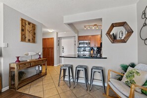 Une cuisine compacte dotée d’appareils électroménagers modernes, une table à manger en bois avec des tabourets, et un coin salon confortable avec un fauteuil et un canapé.