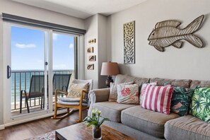 A living room with a sliding glass door leading to a balcony, a sofa with decorative pillows, a rocking chair, and wall art.