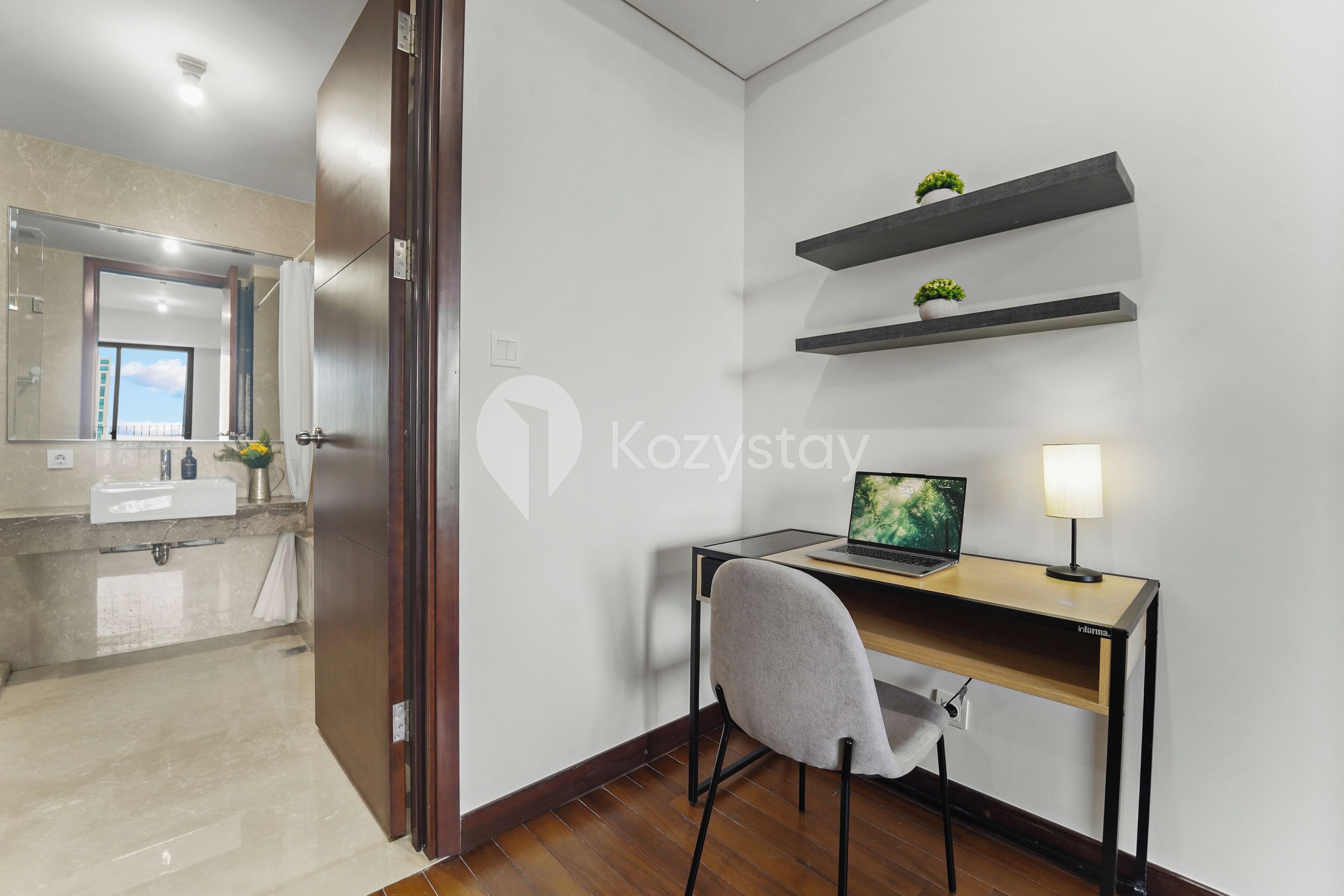 A modern hotel room with a wooden desk, a laptop, and a lamp. There is a bathroom with a mirror and a sink.