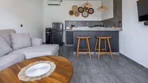 A modern living room with a grey sofa, a wooden coffee table, and a kitchen area with a grey countertop, two wooden stools, and a hanging light fixture. - Oceanview Escape 1BR Villa Bingin, Uluwatu (Bali)