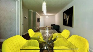 Un coin repas avec une table en verre et quatre chaises jaunes, un luminaire suspendu moderne et une œuvre d’art encadrée au mur.