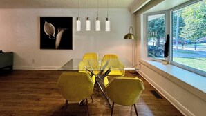Un espace repas moderne avec une table en verre et des chaises jaunes, un luminaire suspendu et une œuvre d’art encadrée au mur.