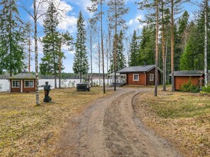 A rustic cabin area with a dirt path leading to a wooden structure, surrounded by tall trees and a body of water. - Louhuranta by Interhome (Karstula)