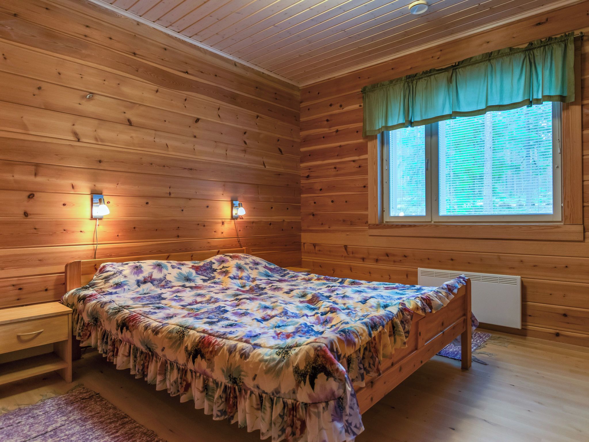 A wooden cabin room with a bed, two wall-mounted lights, a window with blinds, and a radiator.