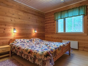A wooden cabin room with a bed, two wall-mounted lights, a window with blinds, and a radiator.